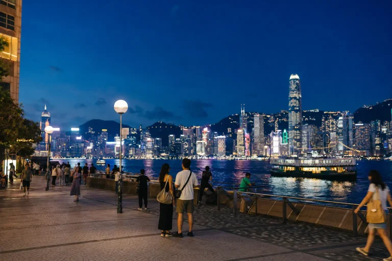 People walking beside Victoria Harbour with the Hong Kong skyline illuminated at night