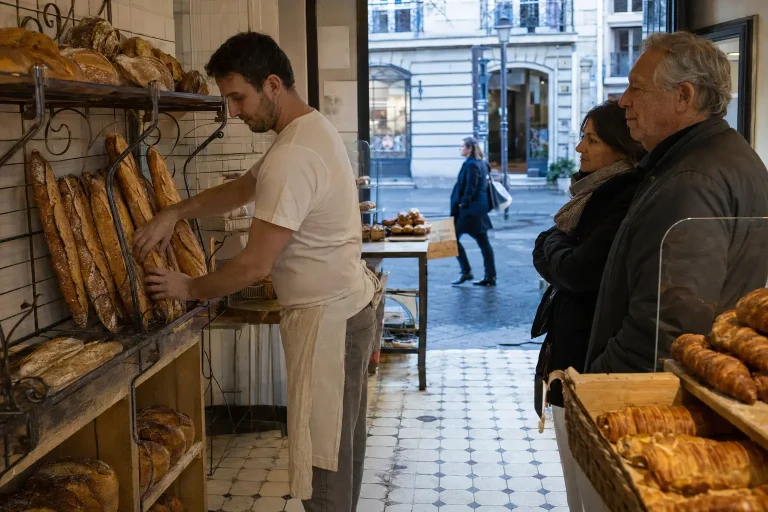 Baker arranging fresh baguettes as customers wait in a neighborhood Paris bakery