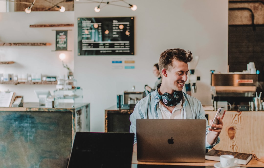 Male at a coffee shop smiling at the phone while working on the computer. — 50+ Best Digital Nomad Jobs to Work From Anywhere