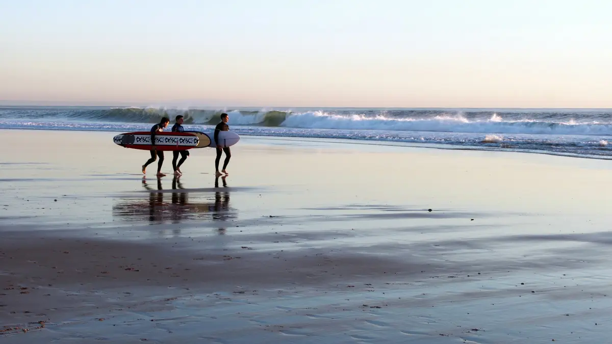 Three surfers in wetsuits carrying boards along the wet sand at Carcavelos Beach near Lisbon