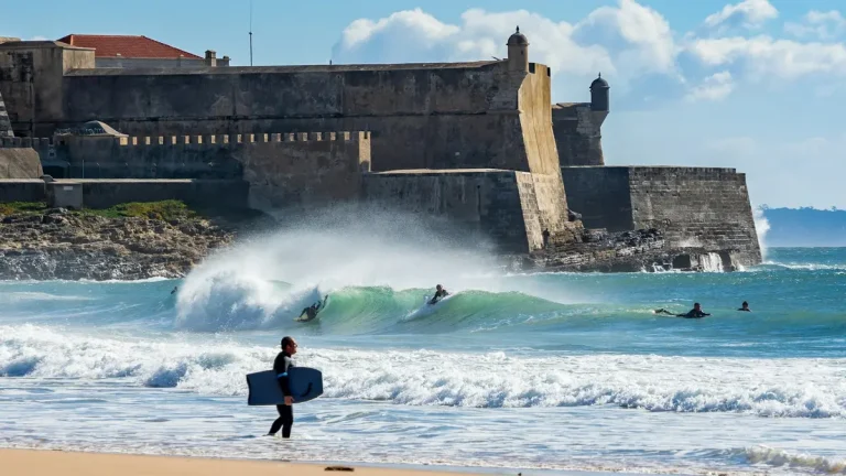 Surfers in the water beside the sandy shore and fort at Carcavelos Beach
