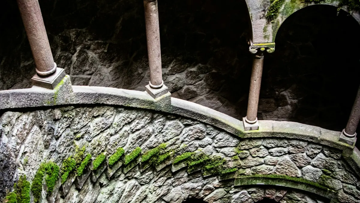 Moss-covered stone walls, columns and arches curving around a spiral well at Quinta da Regaleira in Sintra